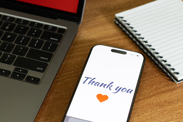 Mobile screen with Thank you message with heart symbol on the wooden table along with the laptop and notes on it.
