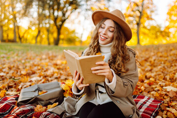 A cute woman in hat sits on soft blanket with book in autumn park. Beautiful woman relaxes in sunny...