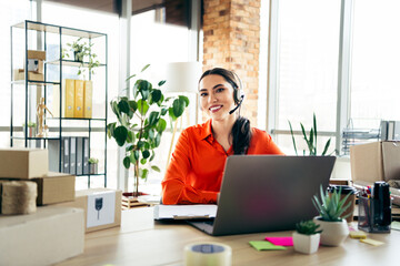 Smiling young businesswoman working with a headset and laptop in a creative workspace with plants and boxes