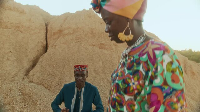 Young Black woman and man wearing colorful ethnic clothes having fun dancing while their friend playing African goblet drum