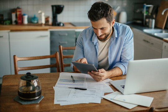 Young man using a digital tablet in the kitchen while going over his bills