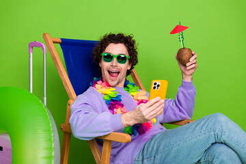 Young man in casual attire sitting on a beach chair with tropical props and phone against a green...