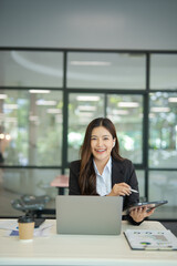 Fototapeta premium Asian businesswoman working at desk using laptop and notebook, talking with client or modern office building.