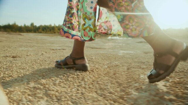 Side view tracking shot of unrecognizable Black woman wearing sandals and patterned dress walking along semi-desert