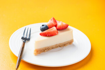 Slice of classical cheesecake with fresh berries topping, served on white plate, yellow background
