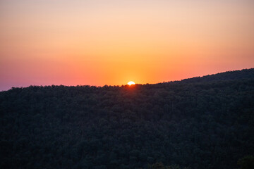 The sun sets behind a forested hill, painting the sky in warm hues of orange and pink as silhouettes embrace the fading light.