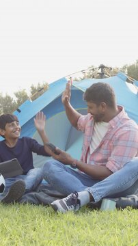 High-fiving Indian father and son sitting at campsite, with blue camping tent and soccer ball