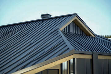Fototapeta premium Modern house roof detail with dark gray corrugated metal panels. Contemporary building exterior design featuring natural wood accents and a chimney under blue sky.
