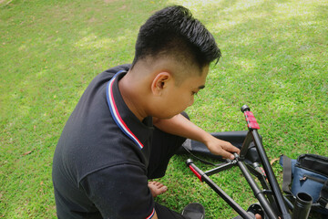 A young man is sitting on a grassy lawn, looking down and adjusting a piece of black Tripod. He is...