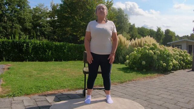 Overweight senior woman standing and sitting in a chair in garden to improve balance, doing squats to strengthen legs and core. Home workout plan for seniors seeking a healthy lifestyle