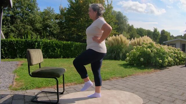 Overweight senior woman in garden lifts legs with help of a chair, strengthening core and improving flexibility. Outdoor home workout plan for seniors seeking a healthy lifestyle