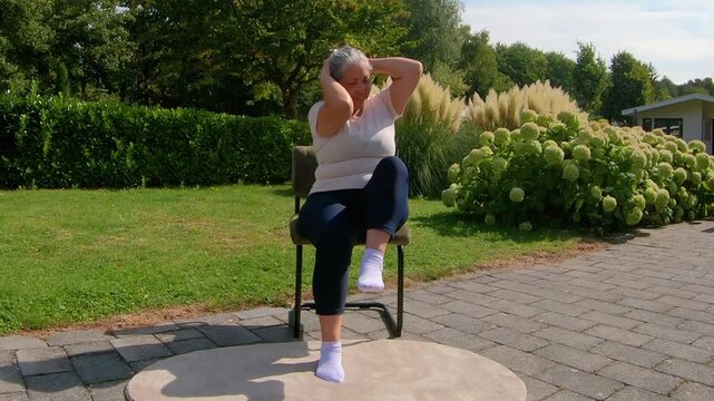 Front view of overweight adult woman strengthening abdomen while sitting in chair in garden, trying to touch elbows and knees. Home workout plan for seniors seeking a healthy lifestyle