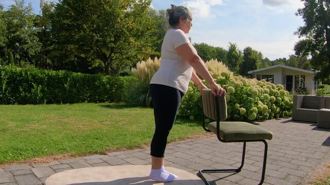 Overweight senior woman lifting one leg while holding onto chair in garden, strengthening to improve flexibility. Home workout plan for seniors seeking a healthy lifestyle