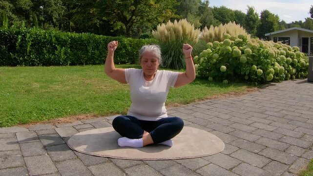 Overweight senior woman strengthening arms while sitting on mat in the garden, opening and closing without weights. Outdoor home workout plan for seniors seeking a healthy lifestyle