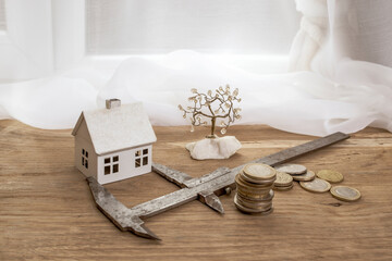 Metal caliper measuring a white model house next to a stack of coins on a wooden desk, construction cost assessment concept