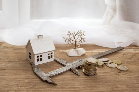 Metal caliper measuring a white model house next to a stack of coins on a wooden desk, construction cost assessment concept - Powered by Adobe