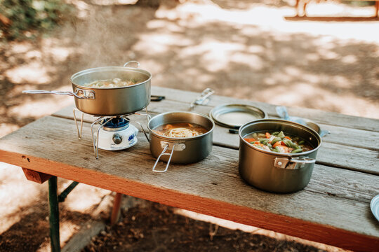 A camping table is set up in the woods, featuring pots of steaming food and plates of pasta topped with shrimp. Nature surrounds the area, creating a cozy outdoor dining experience - Powered by Adobe