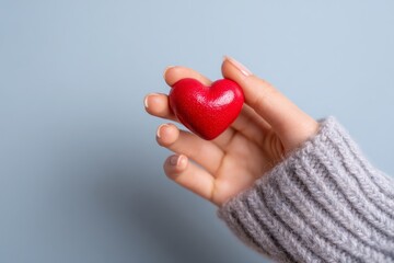 Fototapeta premium A small, vibrant red heart is held gently in a hand wearing a warm gray sweater against a muted backdrop.