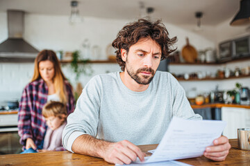 Obraz premium A man reviews documents at a kitchen table while a woman and child are engaged in a separate activity in the background, portraying a family dynamic.
