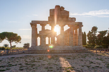 View of the sun shining through the ancient Greek temple ruins, creating a warm glow against the weathered stone, Paestum, Campania, Italy.