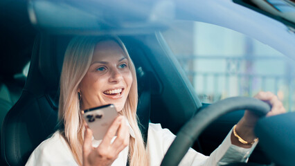 A woman is joyfully driving a car while smiling and using her smartphone in a casual way. A cheerful female happily engaged with her device while driving, showcasing the art of modern multitasking