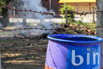 A blue trash can sits outdoors next to a rusty barbed wire fence.