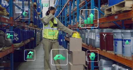 Surveying warehouse worker wearing safety vest holding hardhat shading eyes among drums, with icons