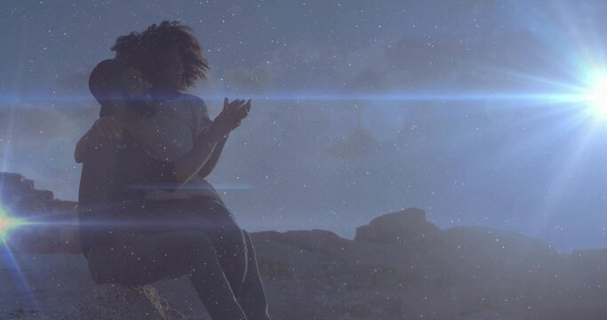 Embracing couple sitting on boulder in desert, with sun casting flares over rocks, copy space - Powered by Adobe