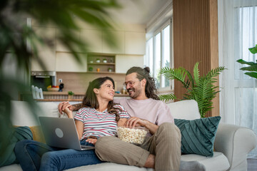 Fototapeta premium Happy couple watching streaming movie on laptop and eating popcorn on sofa at home