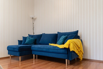 large, beautiful blue sofa in living room.  pillows and yellow plaid on sofa. There is a coffee table with flowers near the sofa, and a floor lamp emits a soft light. Cozy atmosphere in the apartment.