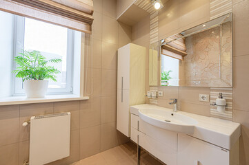 A bright bathroom with beige tiles, a white sink vanity, a mirror, and a window with a plant
