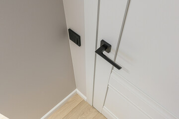 A close-up of a white door with a sleek black handle and a black light switch on a light grey wall,...