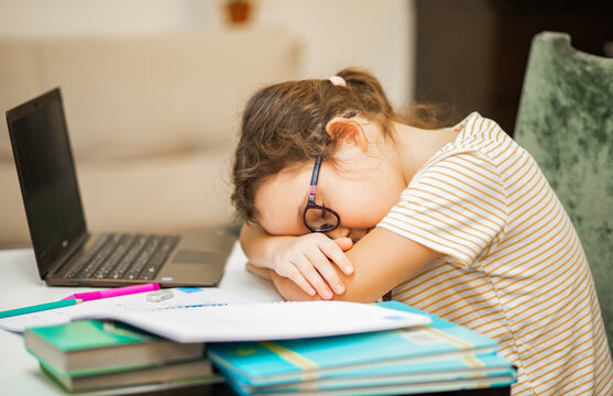 Little cute girl pupil at home making homework. Kid sleeping resting head on books. Child studying online on laptop. Kid education.