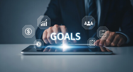 Hands interact with a tablet displaying "GOALS" and related icons: graph, team, checklist, dollar sign.  Illustrates business planning and achievement