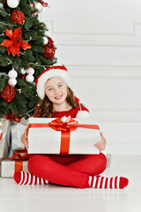 Smiling girl in Santa hat and red pajamas sitting by decorated Christmas tree holding a large white gift box with red ribbon
