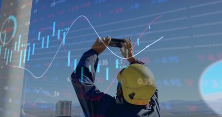 Field worker in coveralls and helmet raising smartphone at site, with financial chart overlay