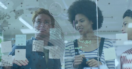 Collaborating colleagues placing sticky notes on glass wall in office, with dots and diagrams