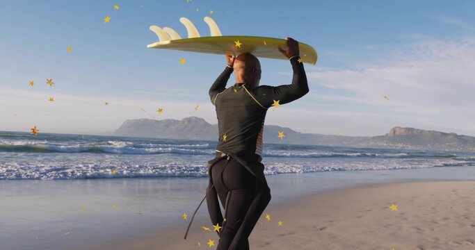 Standing surfer in black wetsuit holding yellow surfboard above head on shore, with ocean waves