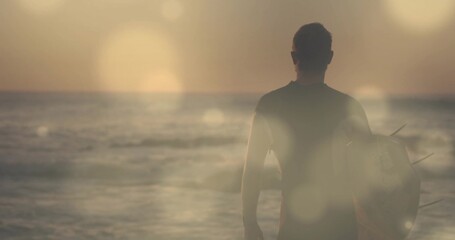 Facing surfer wearing black wetsuit standing on shore holding surfboard, ocean reflecting sunset