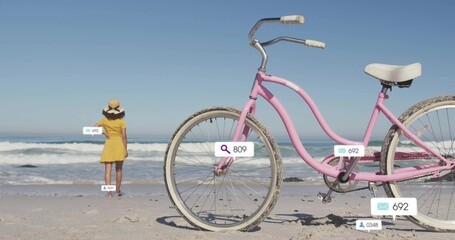 Woman wearing yellow sundress, straw hat standing facing waves on beach, with pastel pink bicycle
