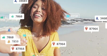 Creator wearing blouse holding smartphone taking selfie on beach with floating social media icons