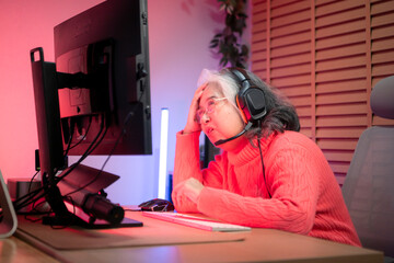 Senior woman gamer wearing headphones and playing on a desktop computer in a colorful gaming setup