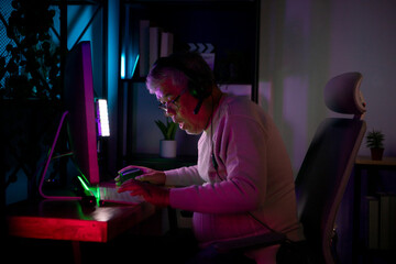 Senior man gamer wearing headphones and playing on a desktop computer in a colorful gaming setup © Wosunan