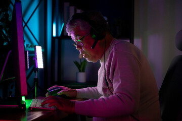 Senior man gamer wearing headphones and playing on a desktop computer in a colorful gaming setup