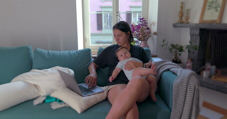 Obraz premium Mother holding baby on her lap while working on a laptop in a cozy living room, managing work and family time simultaneously, focused on her tasks