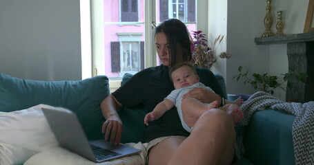 Mother holding her baby while working on a laptop at home, balancing childcare and remote work in a...