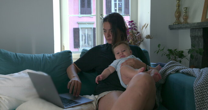 Mother holding her baby while working on a laptop at home, balancing childcare and remote work in a cozy living room setting