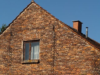 Close-up of an old rustic brick house facade with gable roof, weathered window, and chimney against clear blue sky, showing vintage rural architecture and textured brickwork