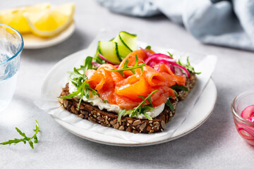 Salmon toast with fresh cucumber, pickled onion, cream cheese and arugula salad on a white plate. Close up.