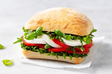 Ciabatta sandwich with mozzarella, tomatoes, pesto sauce and fresh basil on a parchment paper. Light grey background. Close up.